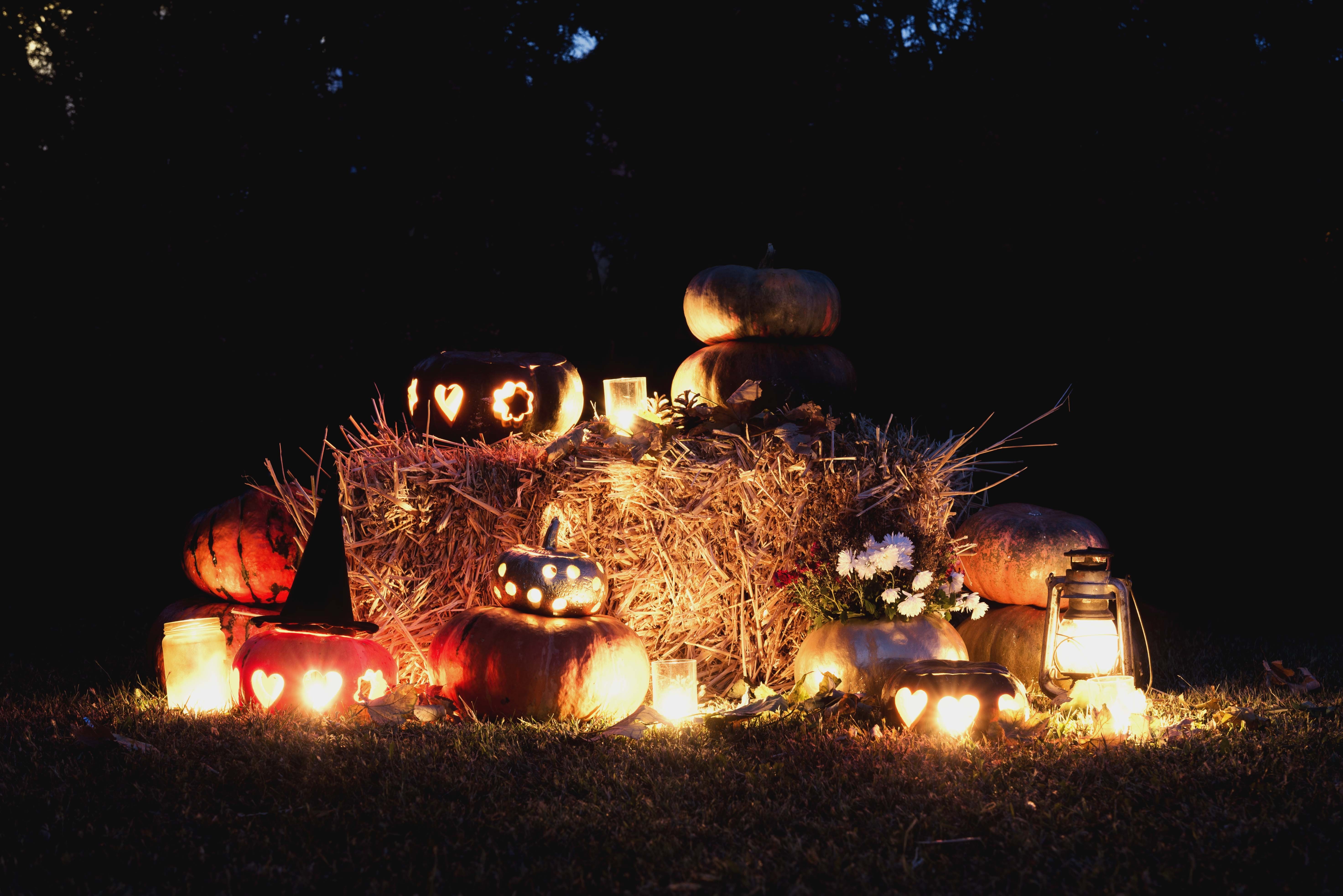 Carved pumpkins with glowing candles sit on hay bales and grass, surrounded by lanterns and flowers on a dark autumn night.
