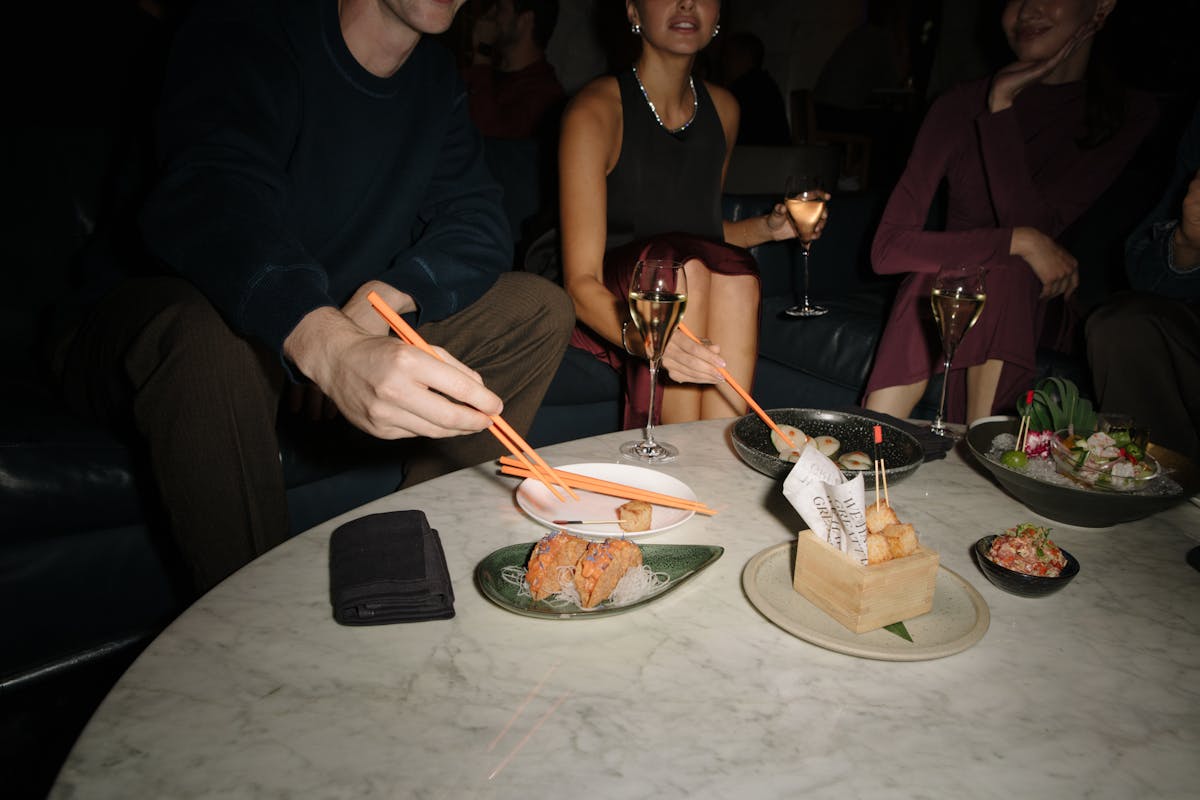 Three friends sitting at a low white table using orange chopsticks to pick up food from plates