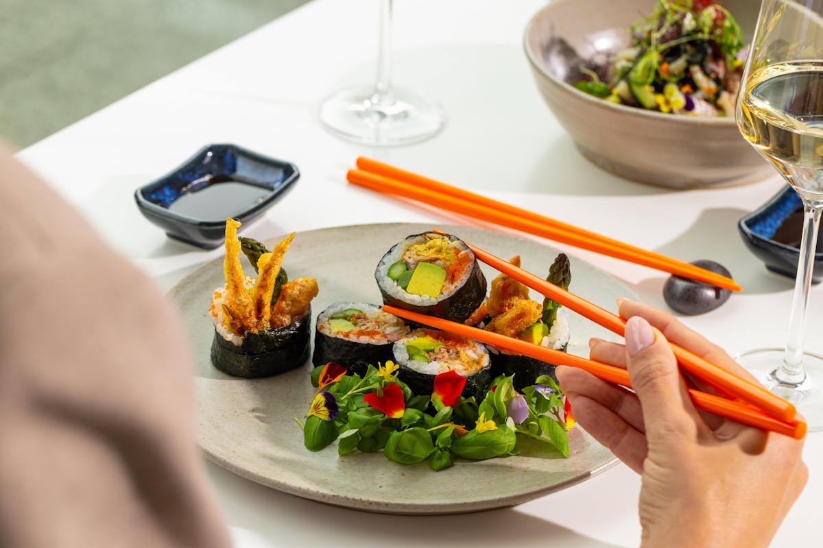 Person holding up a sushi roll from a plate of rolls and salad, using orange chopsticks