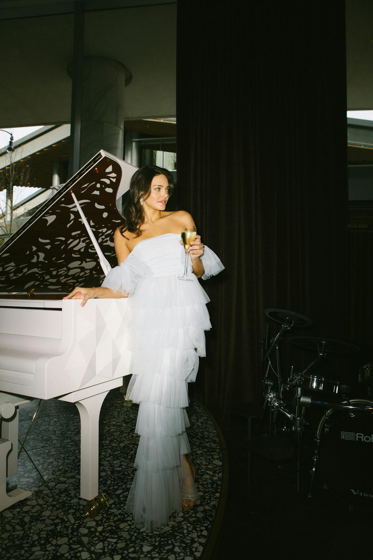 Woman in formal white dress standing next to piano holding glass of champagne
