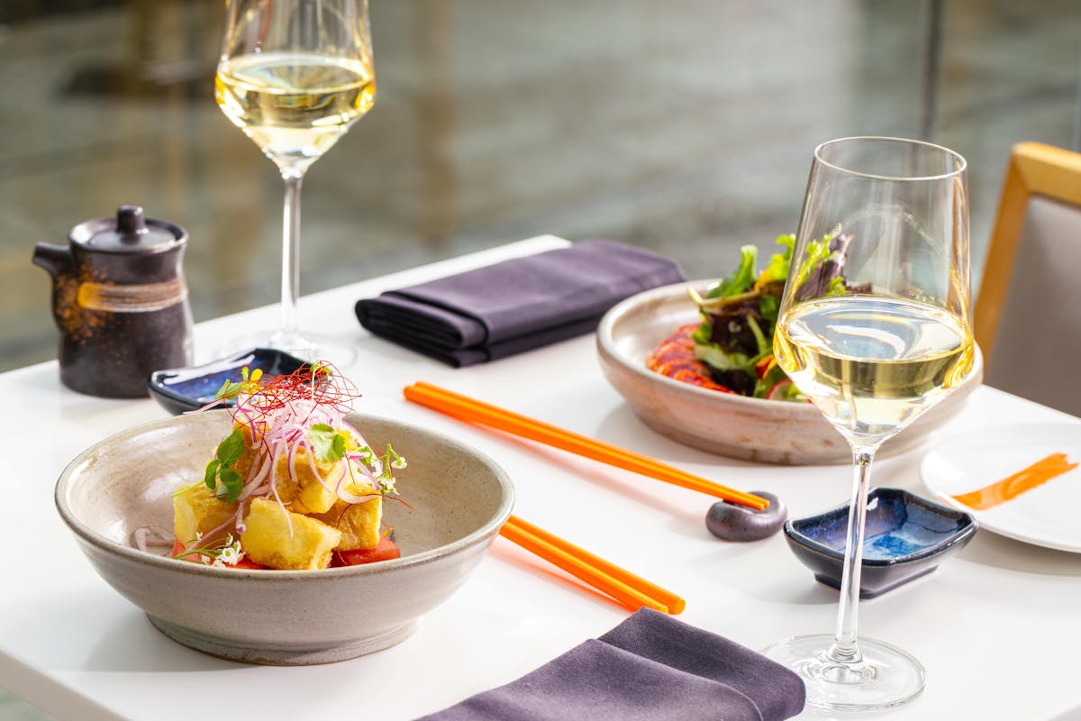 A bowl of tofu salad on a white table, a bowl of lobter salad, two glasses of wine and orange chopsticks on a white table in a lounge in Vancouver