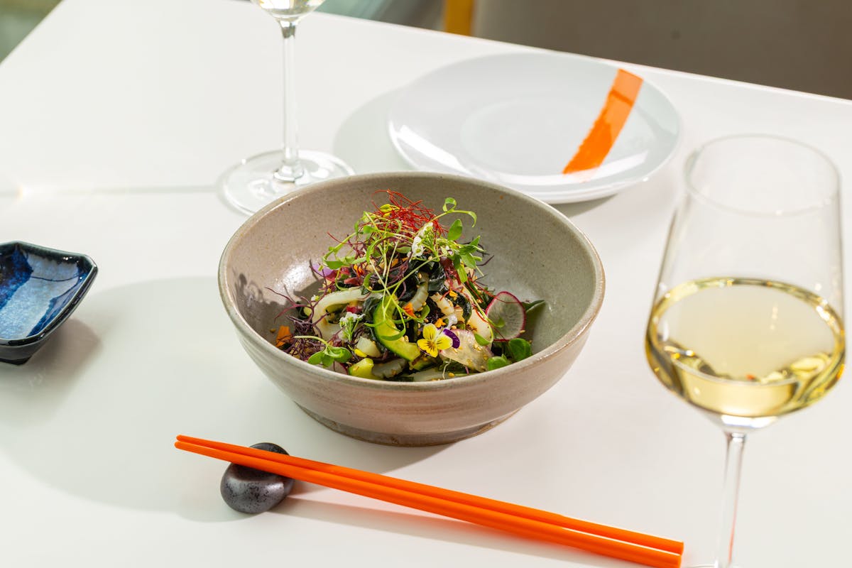 A bowl of salad on a white table next to a glass of wine, orange chopsticks and a white plate with an orange stripe