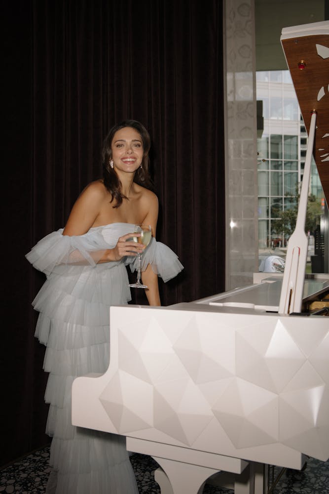Woman in formal white dress standing at a white piano holding glass of champagne