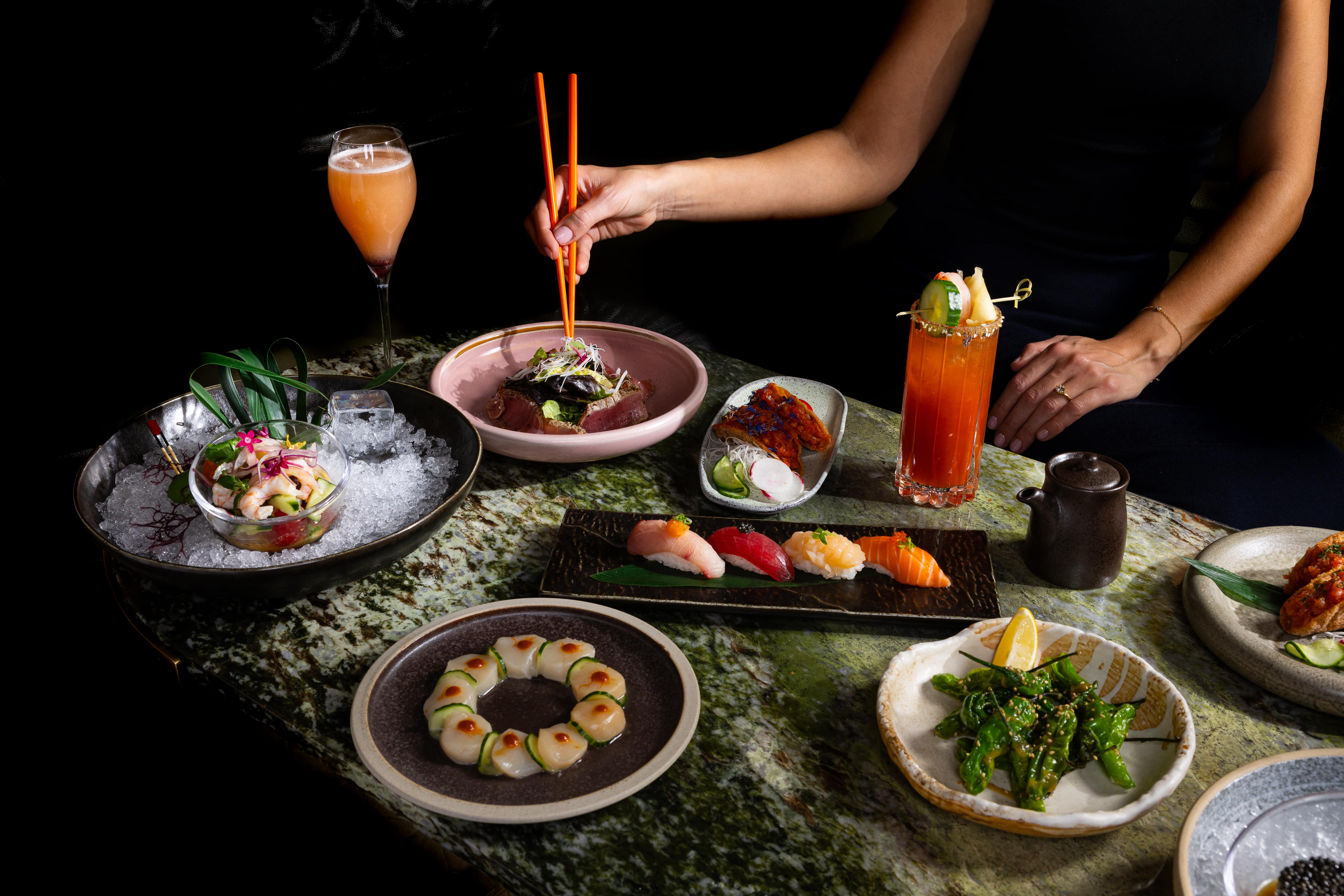A woman sitting at a table with plates of sushi and cocktails, holding orange chopsticks