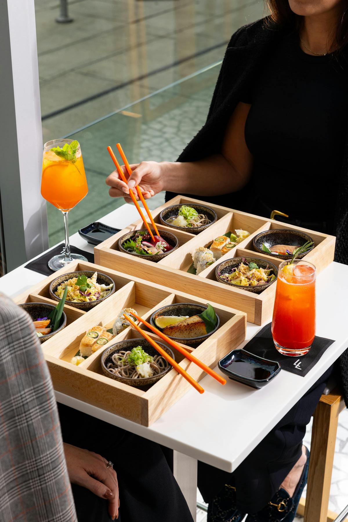 Two people sitting at a table with large wooden bento boxes filled with food, non-alcoholic orange cocktails and orange chopsticks