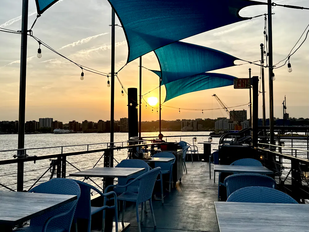 Guests eating at outdoor tables on the deck at Frying Pan NYC as a boat passes on the Hudson.