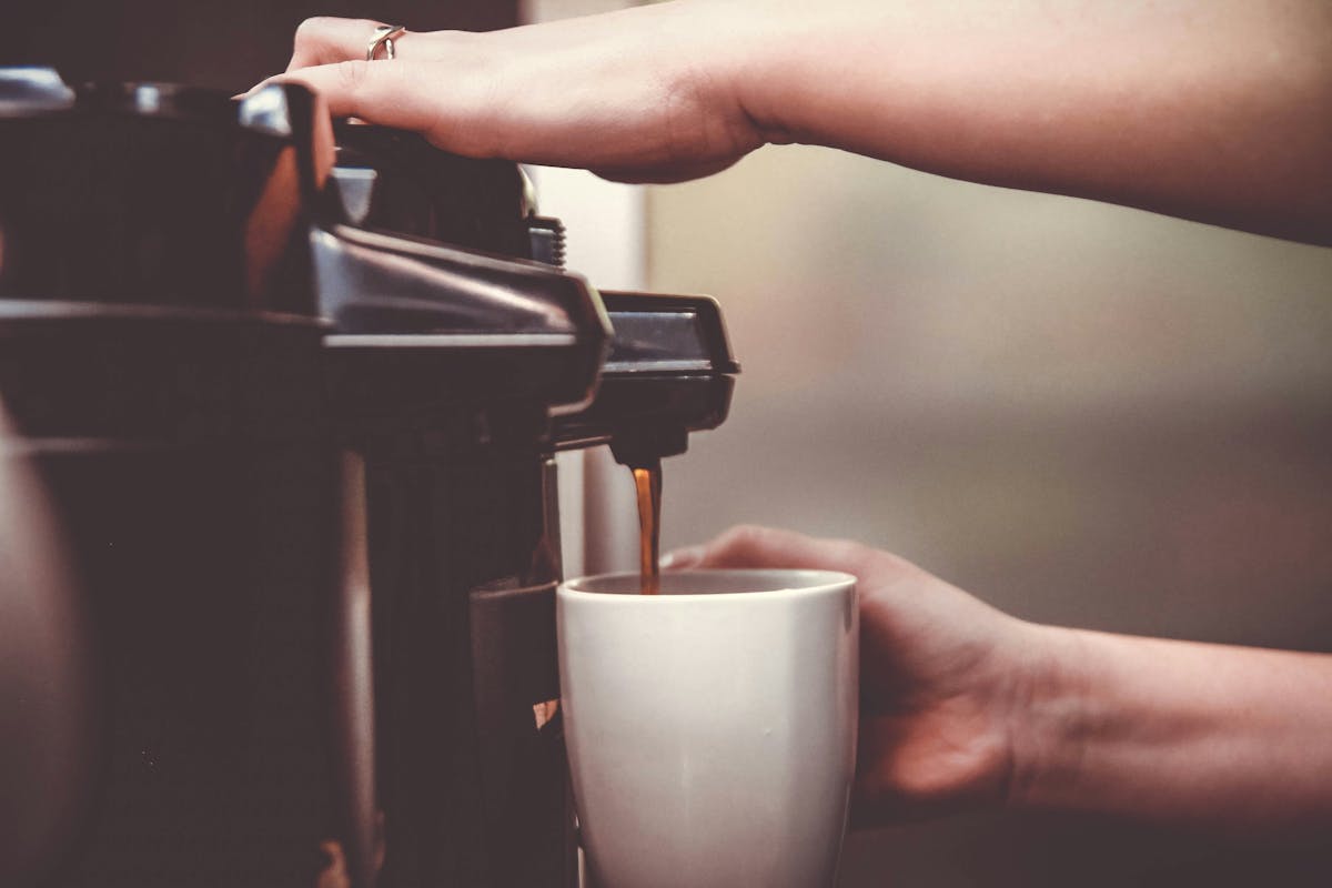 A person pouring freshly brewed coffee from a pot into a white cup on a wooden table