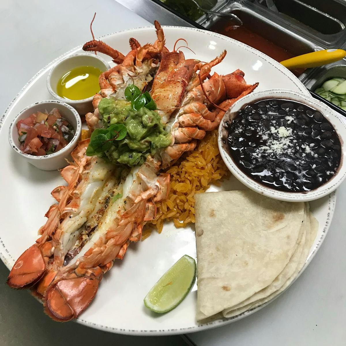 a plate of a lobster tail butterflied with a small bowl of beans, a folded tortilla, a slice of lemon and two sides of salsa
