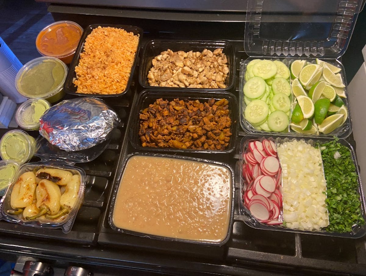 Various trays of different foods, including, slices of radishes, sliced cucumbers, limes, mashed beans, slices of taco meat and chicken, rice and two different salsas in two separate containers