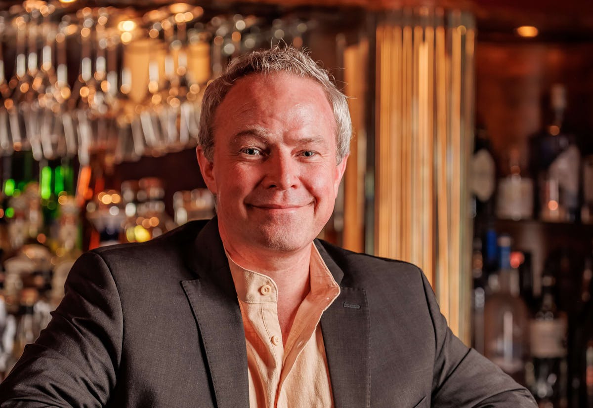 A man seated at a bar, holding a drink, appears relaxed and engaged in conversation