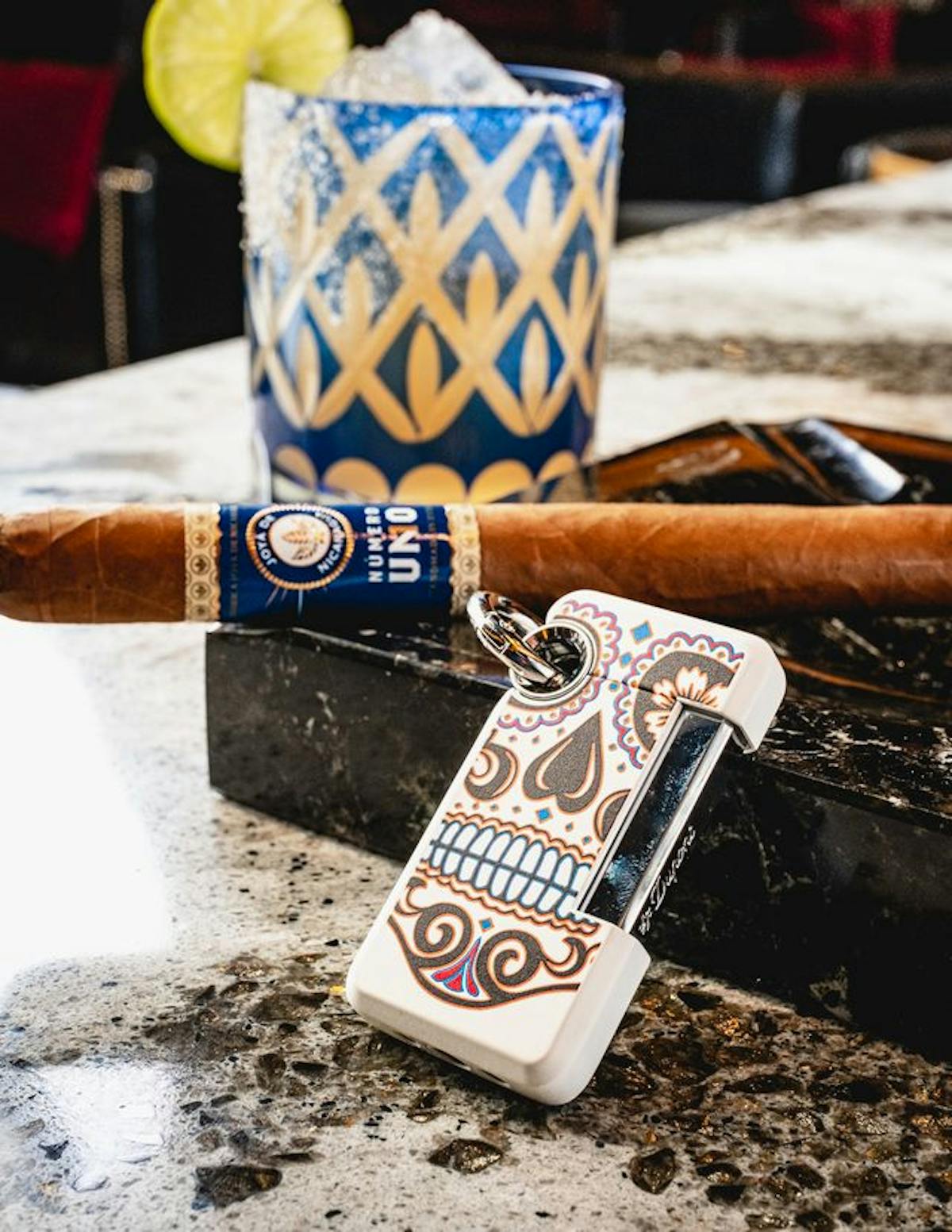 A vibrant photo featuring a cocktail and cigar setting. In the foreground, there's a premium cigar with a blue and gold label, resting beside an intricately patterned, white lighter with paisley designs. Behind these items is a handcrafted cocktail in a blue and white diamond-patterned glass, garnished with a lime wedge. The scene suggests an atmosphere of luxury and relaxation, typical of a high-end cocktail lounge.