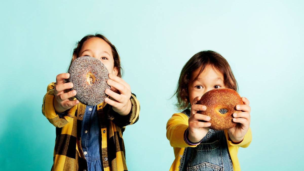 daisy and lulu kostow holding bagels in front of their faces