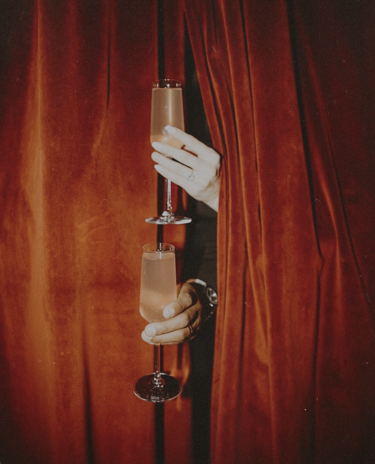 Hands holding champagne flutes through deep red velvet curtains inside Wax Rabbit speakeasy with moody lighting and intimate nightlife atmosphere.