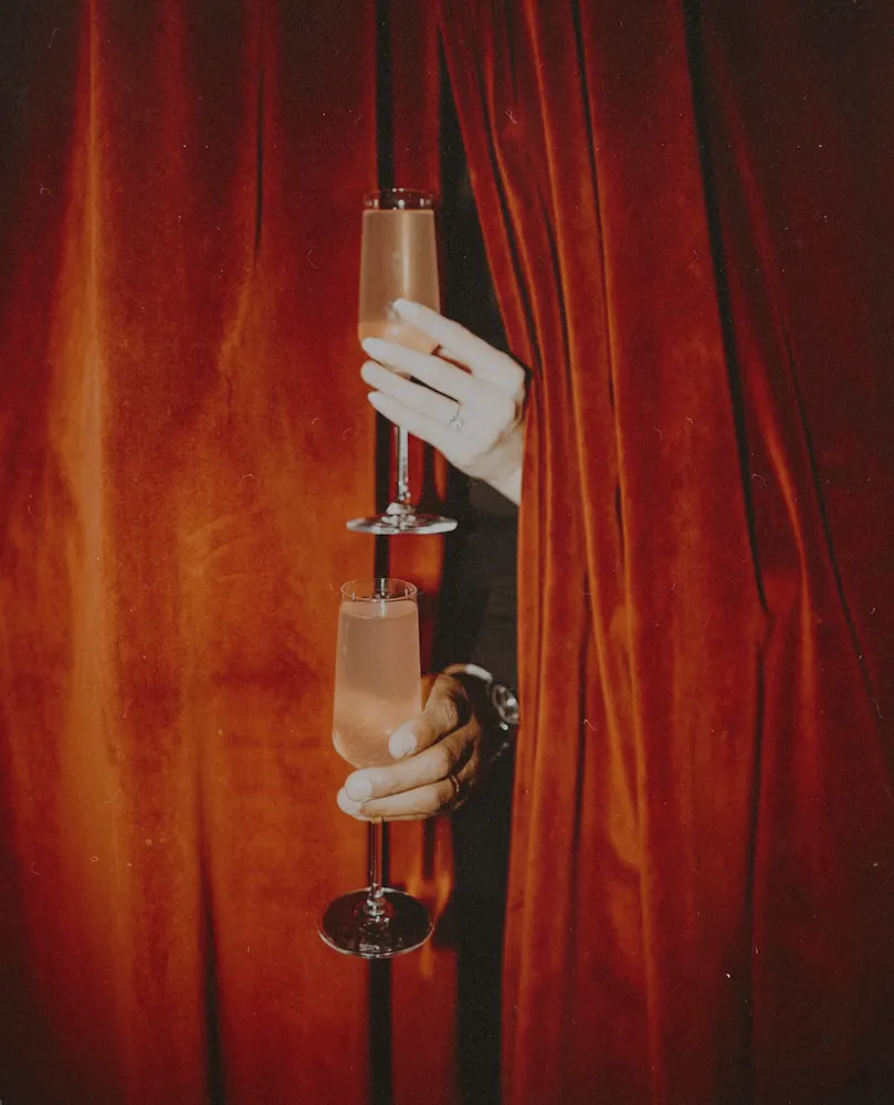 Hands holding champagne flutes through deep red velvet curtains inside Wax Rabbit speakeasy with moody lighting and intimate nightlife atmosphere.