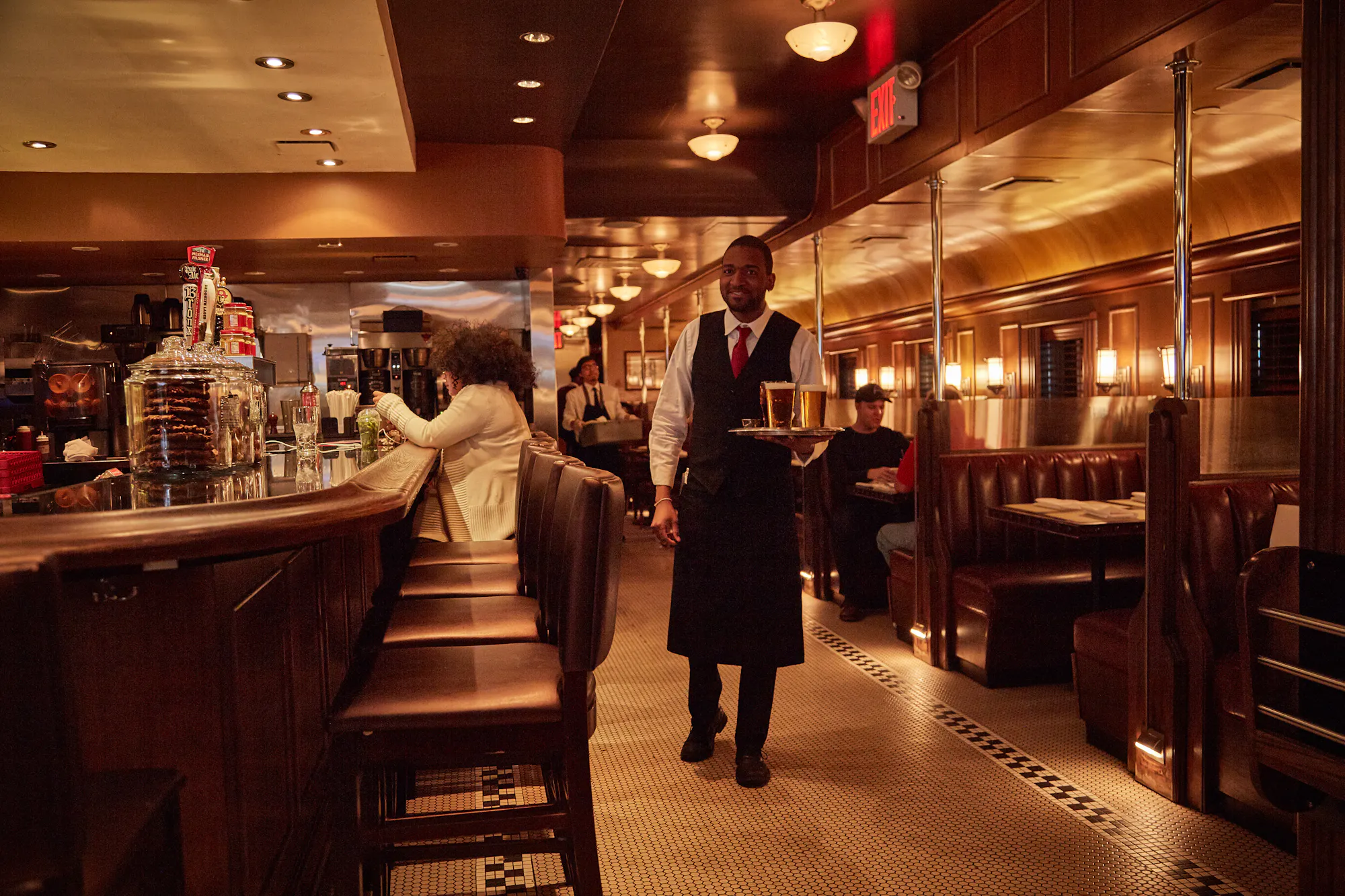 The mahogany and brass dining room at Brooklyn Diner at night, a server carrying a tray past the booths