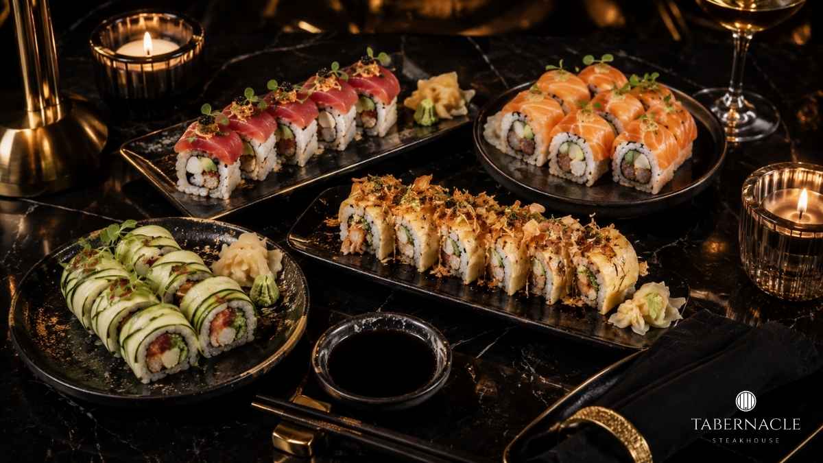 Four sushi roll varieties on black slate plates with soy sauce and candles