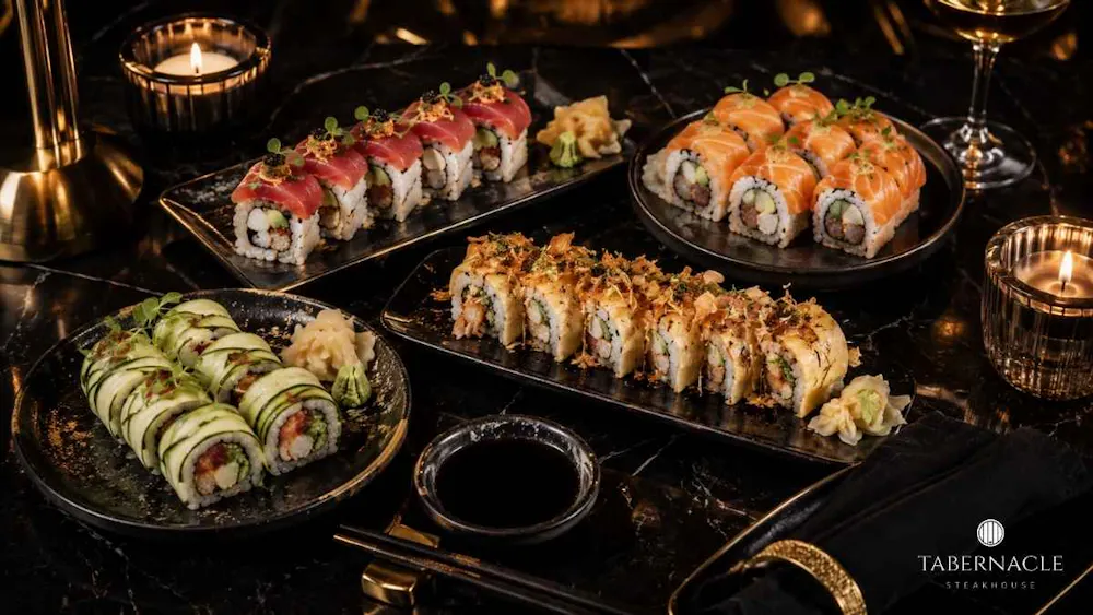 Four sushi roll varieties on black slate plates with soy sauce and candles