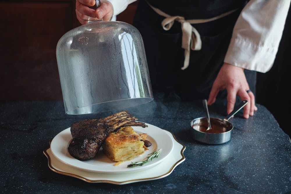 Herb-crusted rack of lamb presented by our Head Chef, Rodrigo, with boulanger-style potatoes and a rosemary jus. Steam can be seen gently rising from the glass cloche covering the dish.