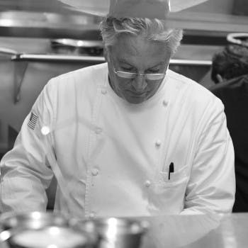 Chef David Slay in a white uniform and glasses looks down while preparing food in a kitchen. The chef has gray hair and a pen in his pocket. The background shows stainless steel surfaces and another person slightly out of focus.