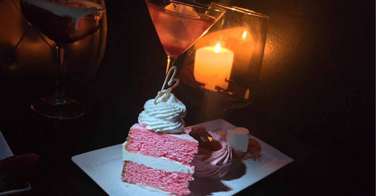 A slice of pink champagne cake and a pink cocktail on a candlelit table at Better Than Sex - A Dessert Restaurant