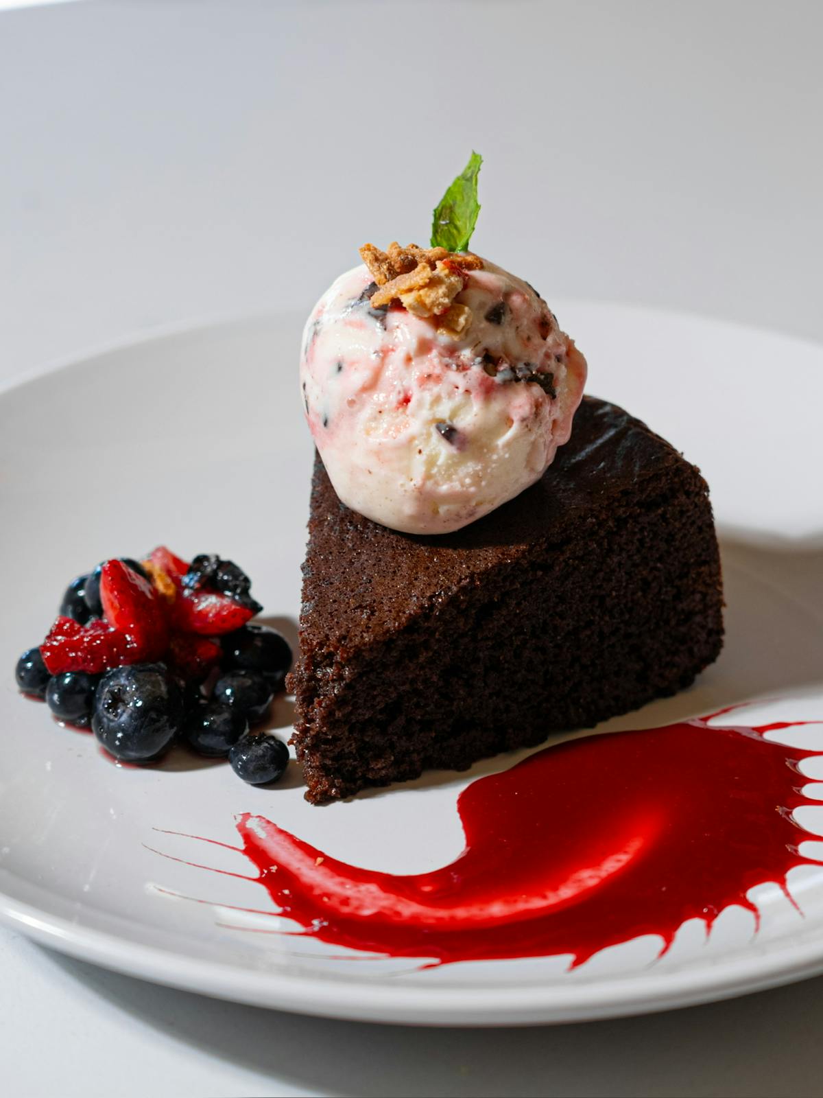 A slice of chocolate cake topped with ice cream and fresh berries, elegantly presented on a white plate.