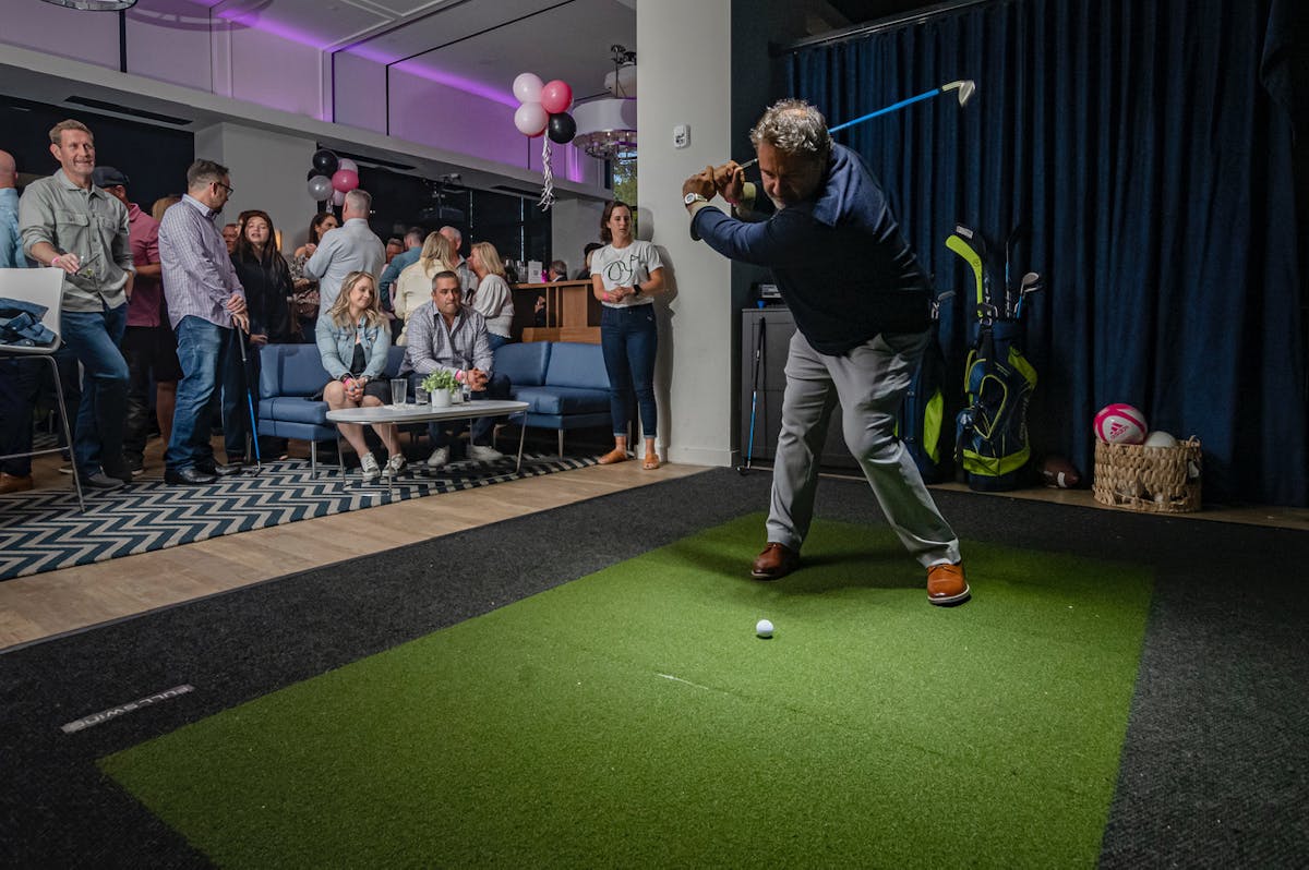 A man swings a golf club, striking a golf ball during a lively outdoor party gathering.