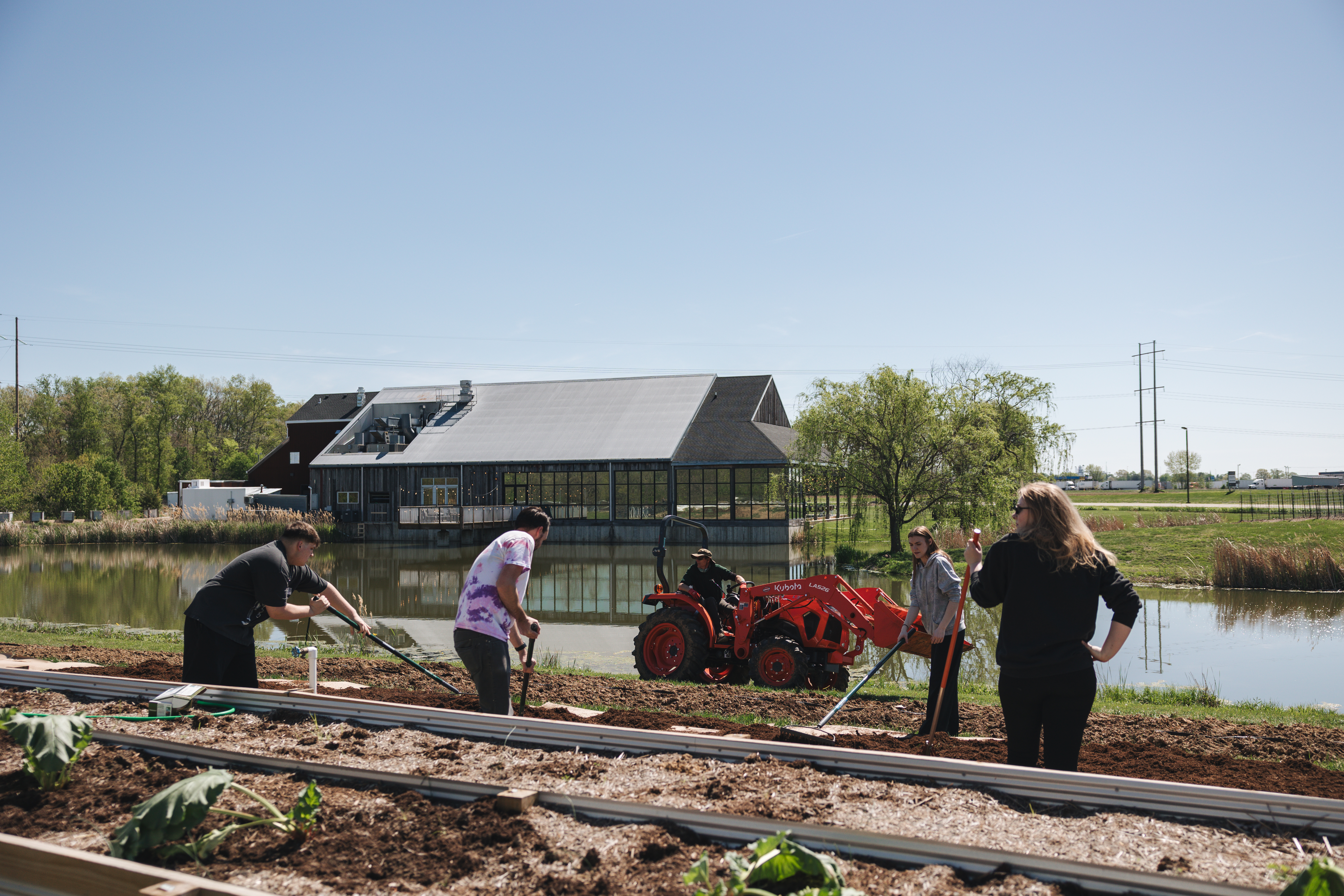 A wide view of the Firefly Farm grounds with the team planting rows near the pond and tractor on Farm Day