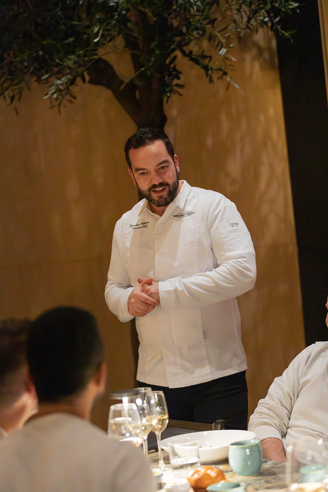  A man in a white shirt stands at a table surrounded by other individuals engaged in conversation
