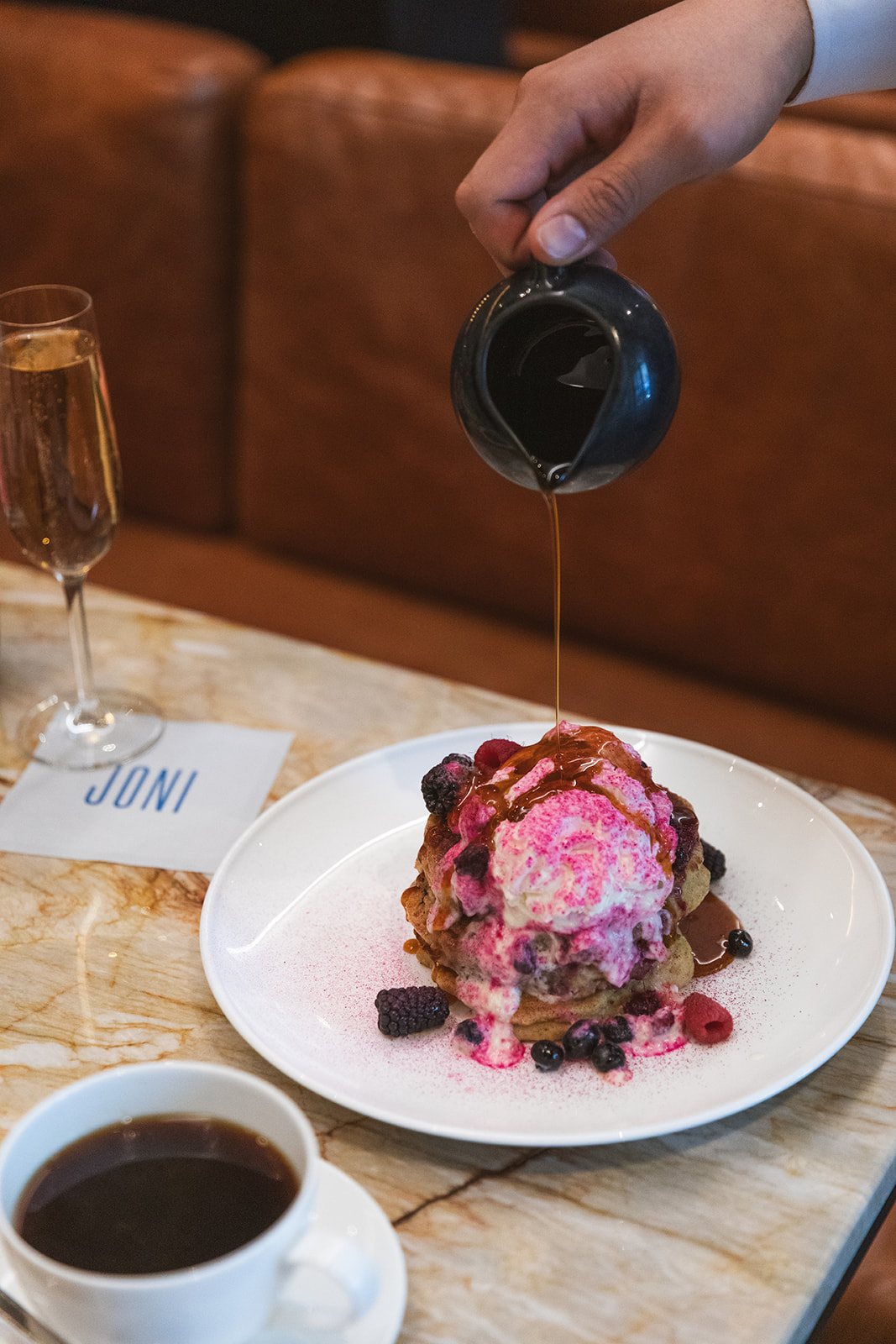 A person drizzling syrup over a stack of fluffy pancakes on a white plate, creating a delicious breakfast scene