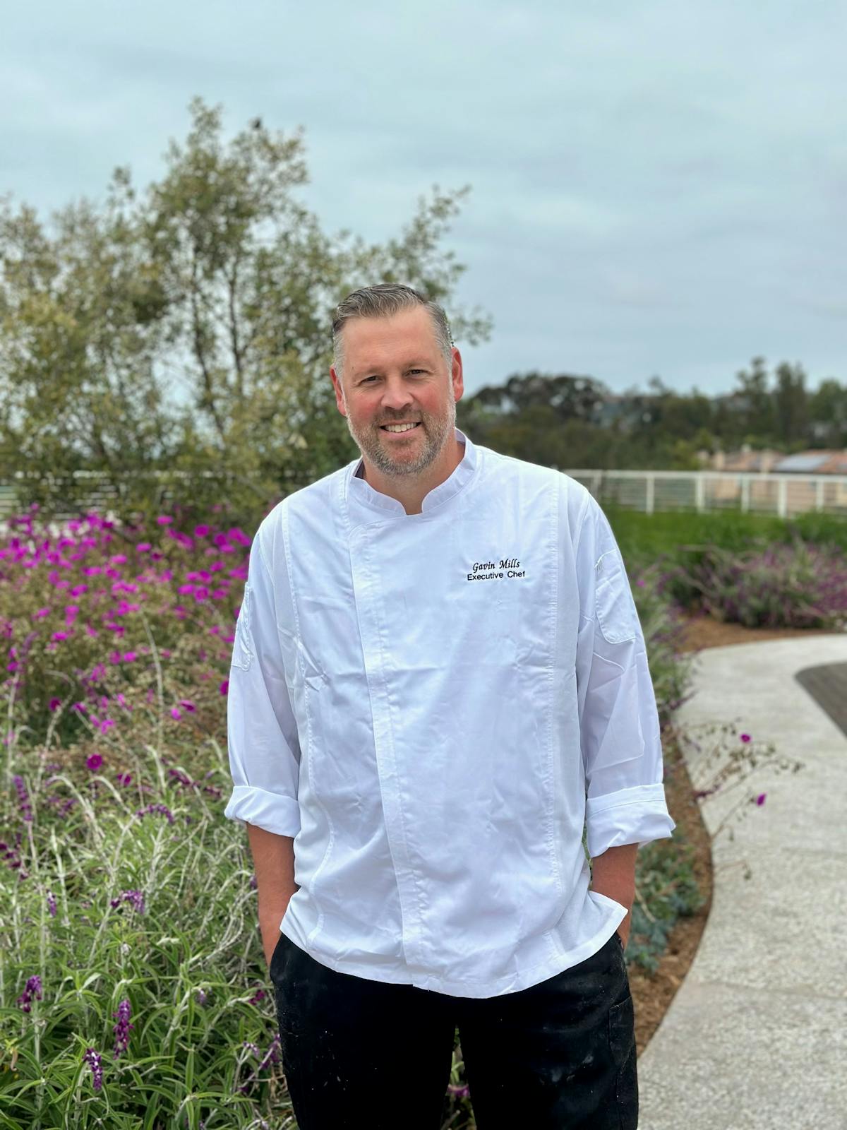A chef in a white chef's coat in front of a field of purple wild flowers