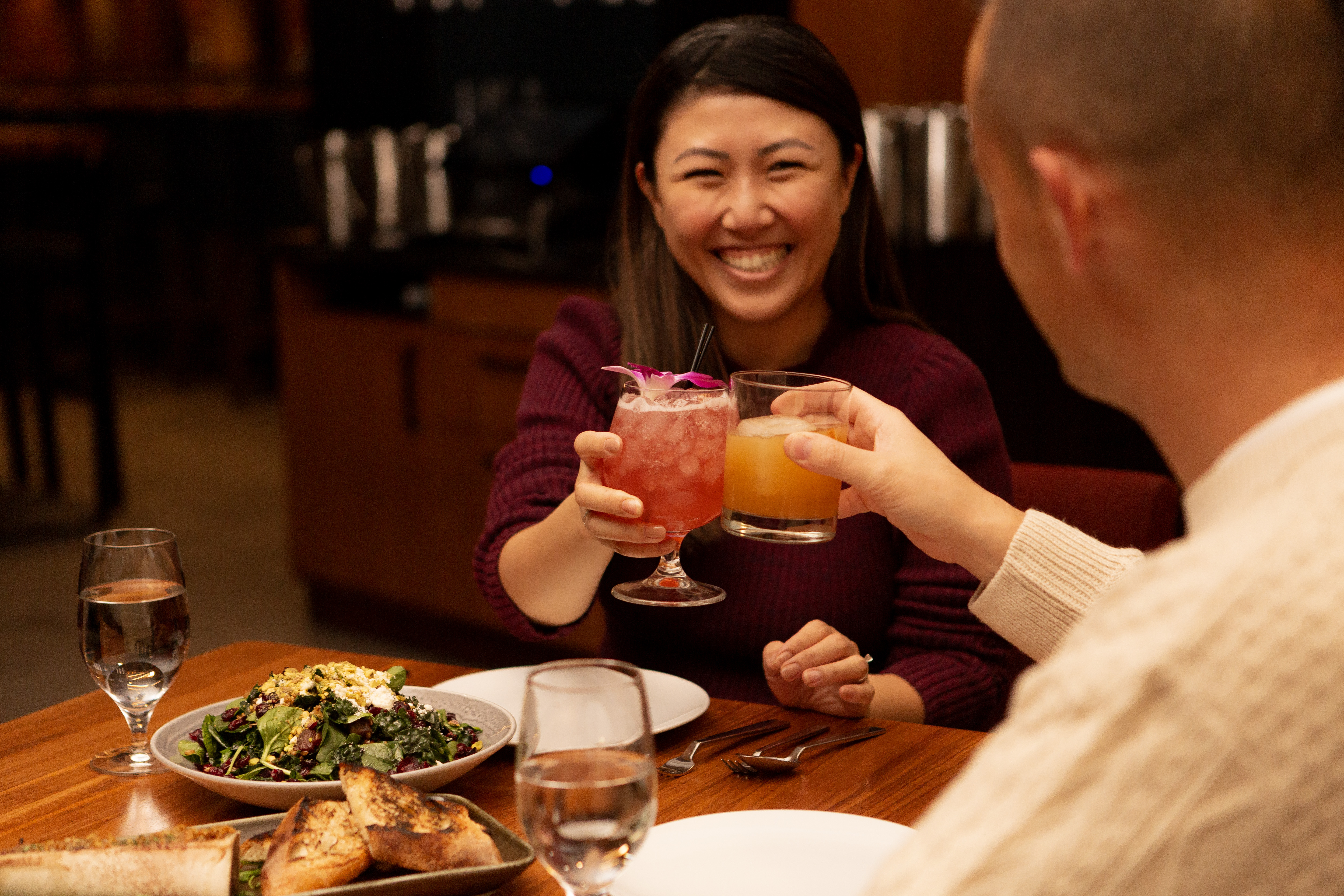 A woman and a man cheering drinks with a salad and bread on a table.