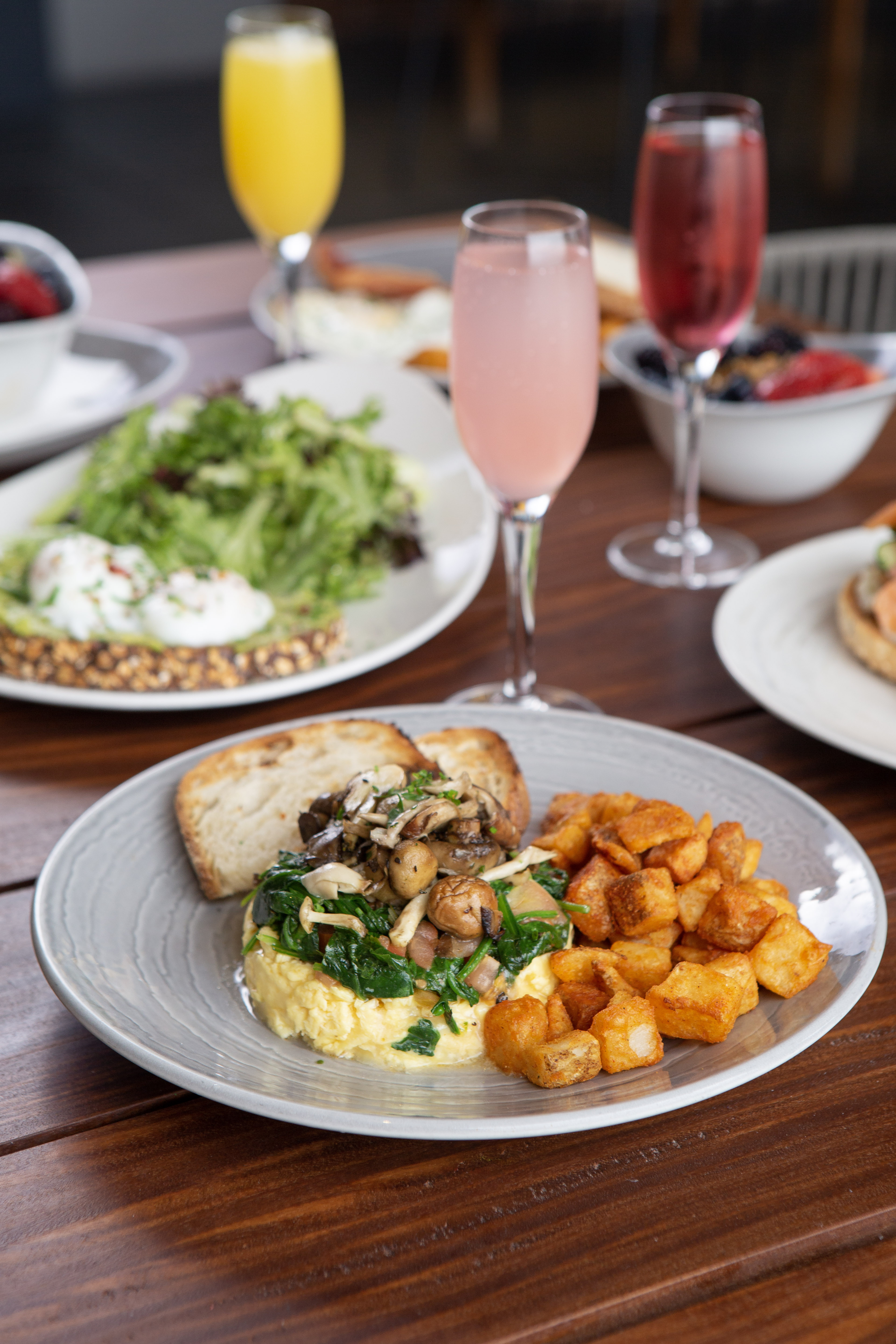 Close up of mushroom egg scramble, toast, and potatoes. Background can see avocado toast with two eggs and the stem of a champagne glass.