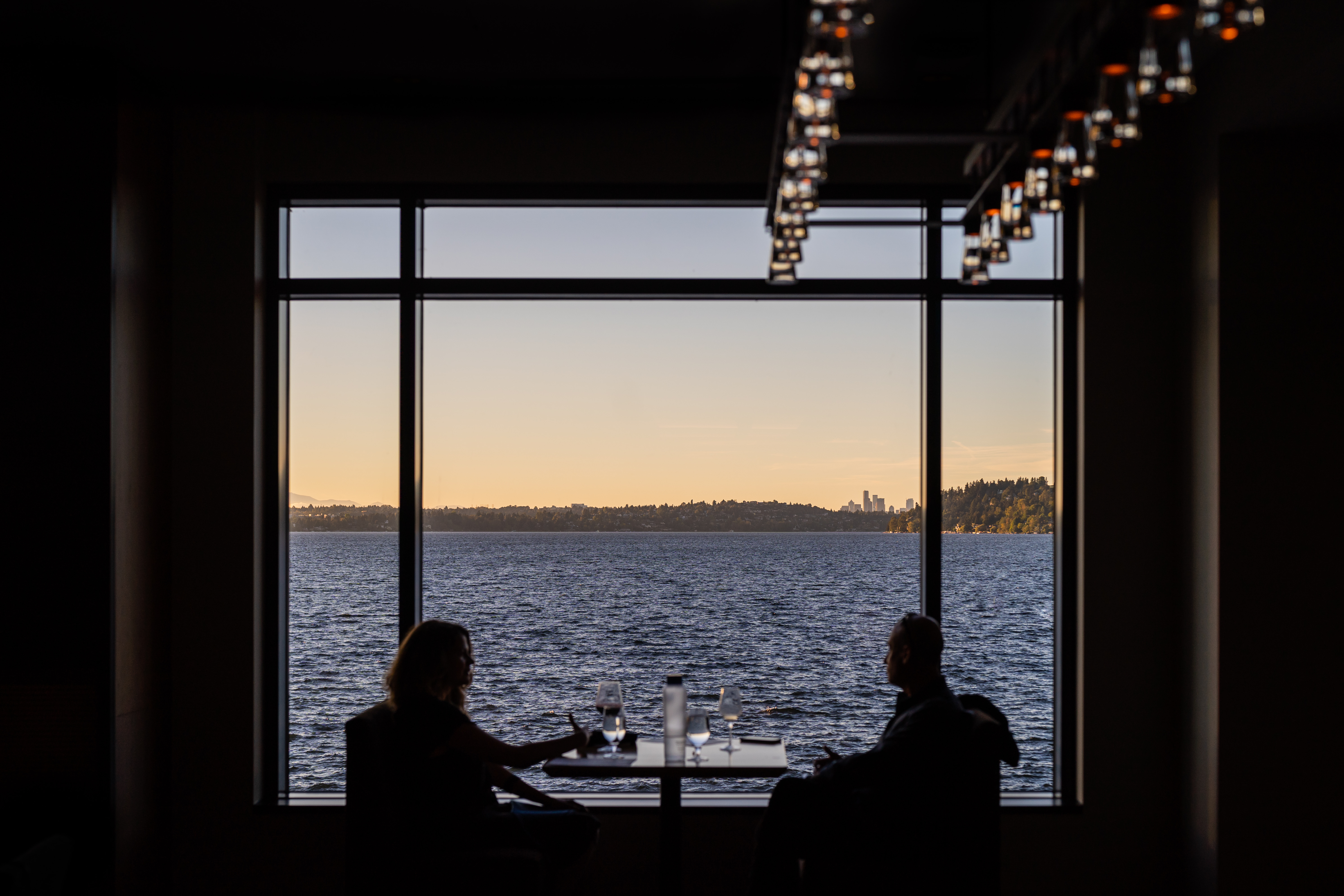 two people in shadow sitting in front of a large window with the sunset and lake Washington in view. Seattle skyline in the distance along with hills.
