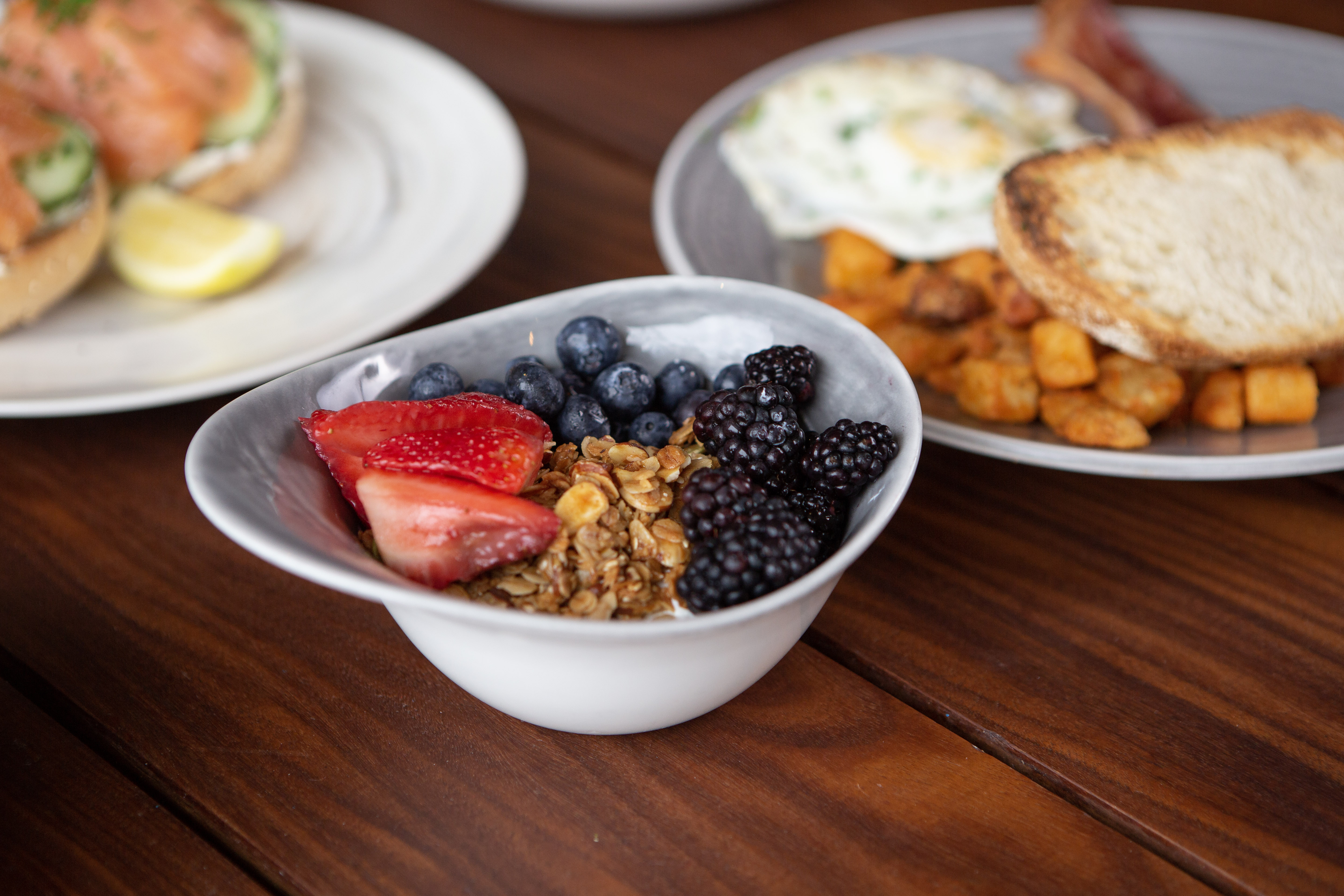 A bowl of granola, blackberries, strawberries, and blueberries. In the background are salmon locks and eggs, potatoes, and toast.