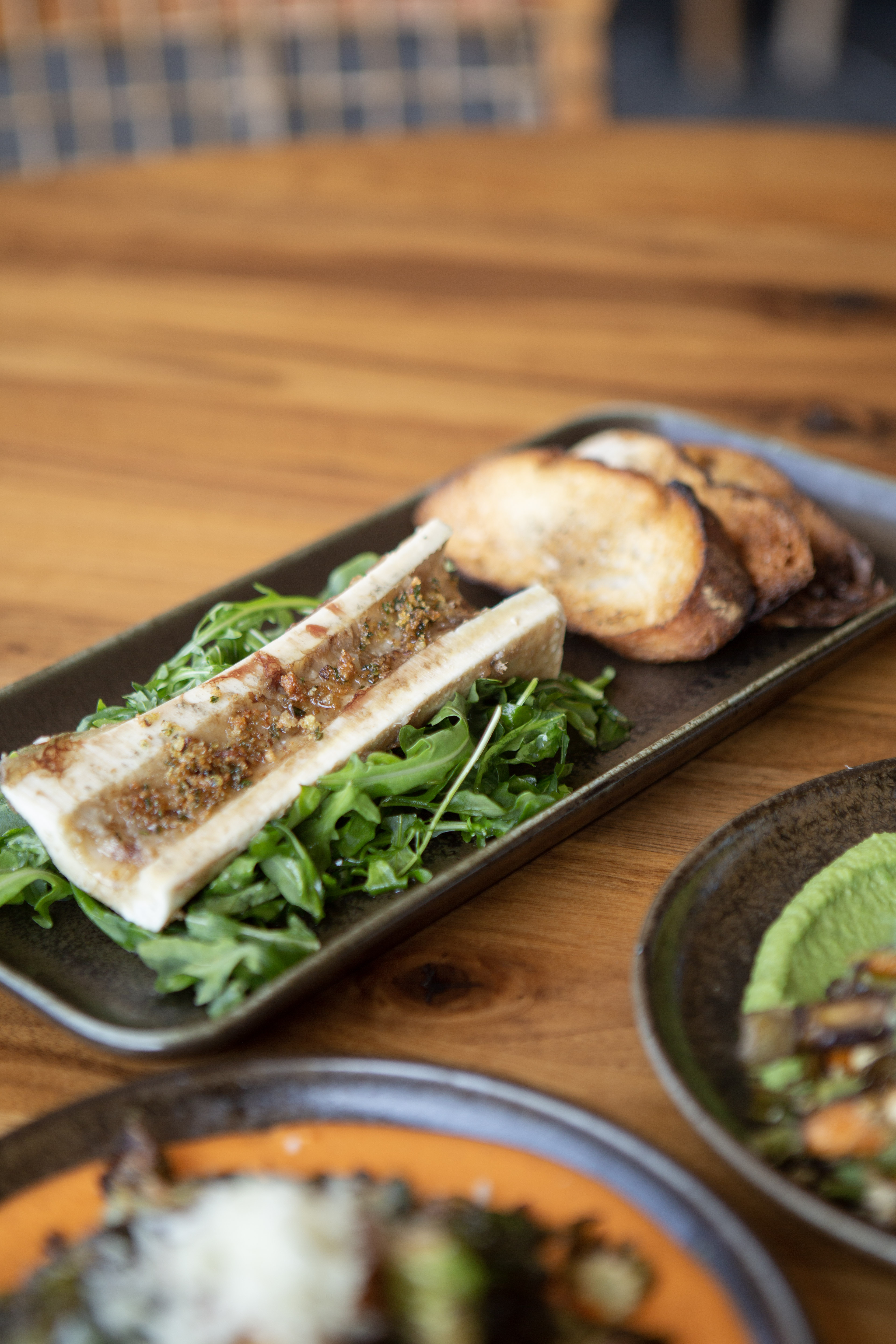 a plate of food on a wooden table of bone marrow, arugula and two pieces of bread.