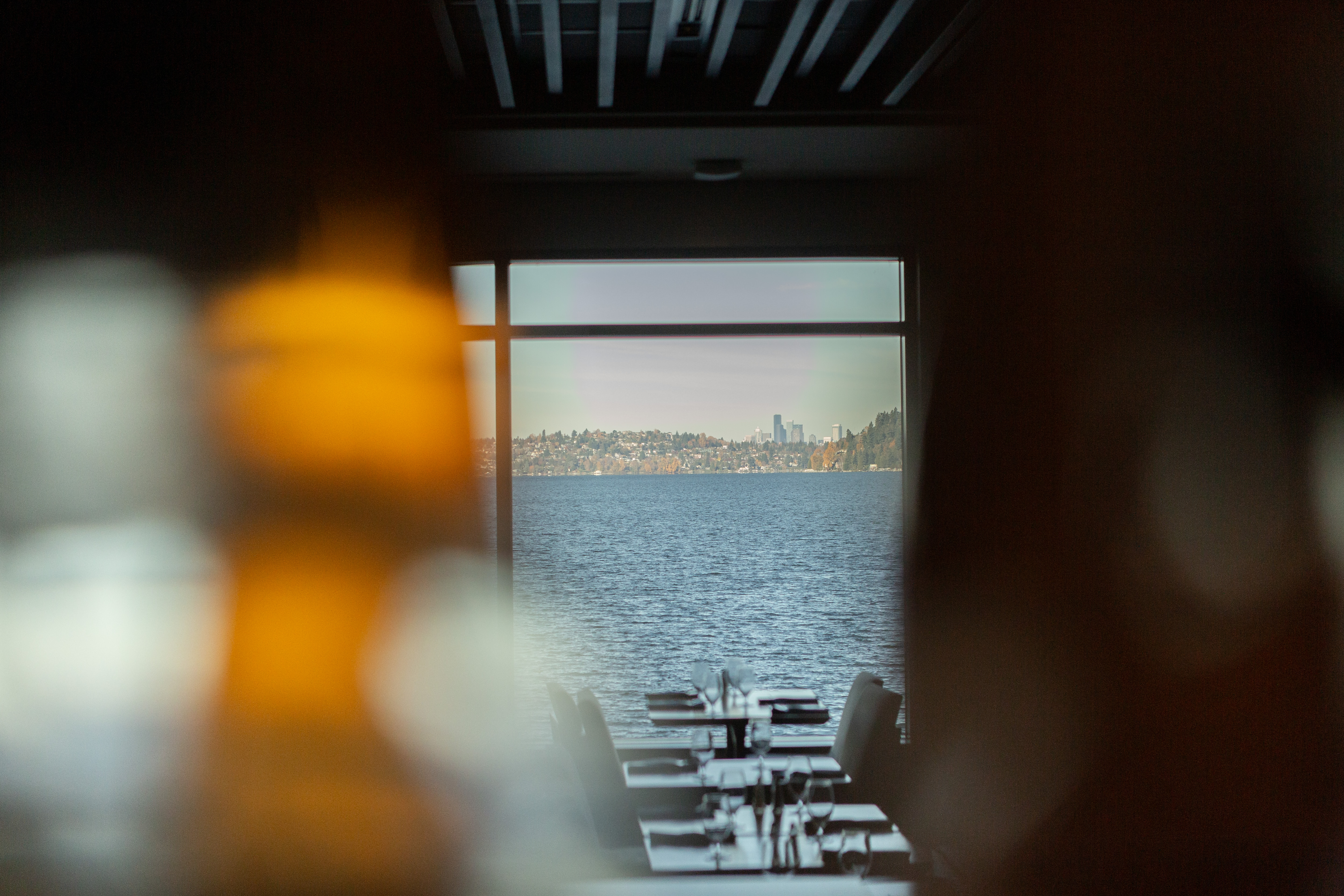 View of interior restaurant with empty tables looking out a window featuring a view of Lake Washington and Seattle skyline. Two bottles blurred in the view.