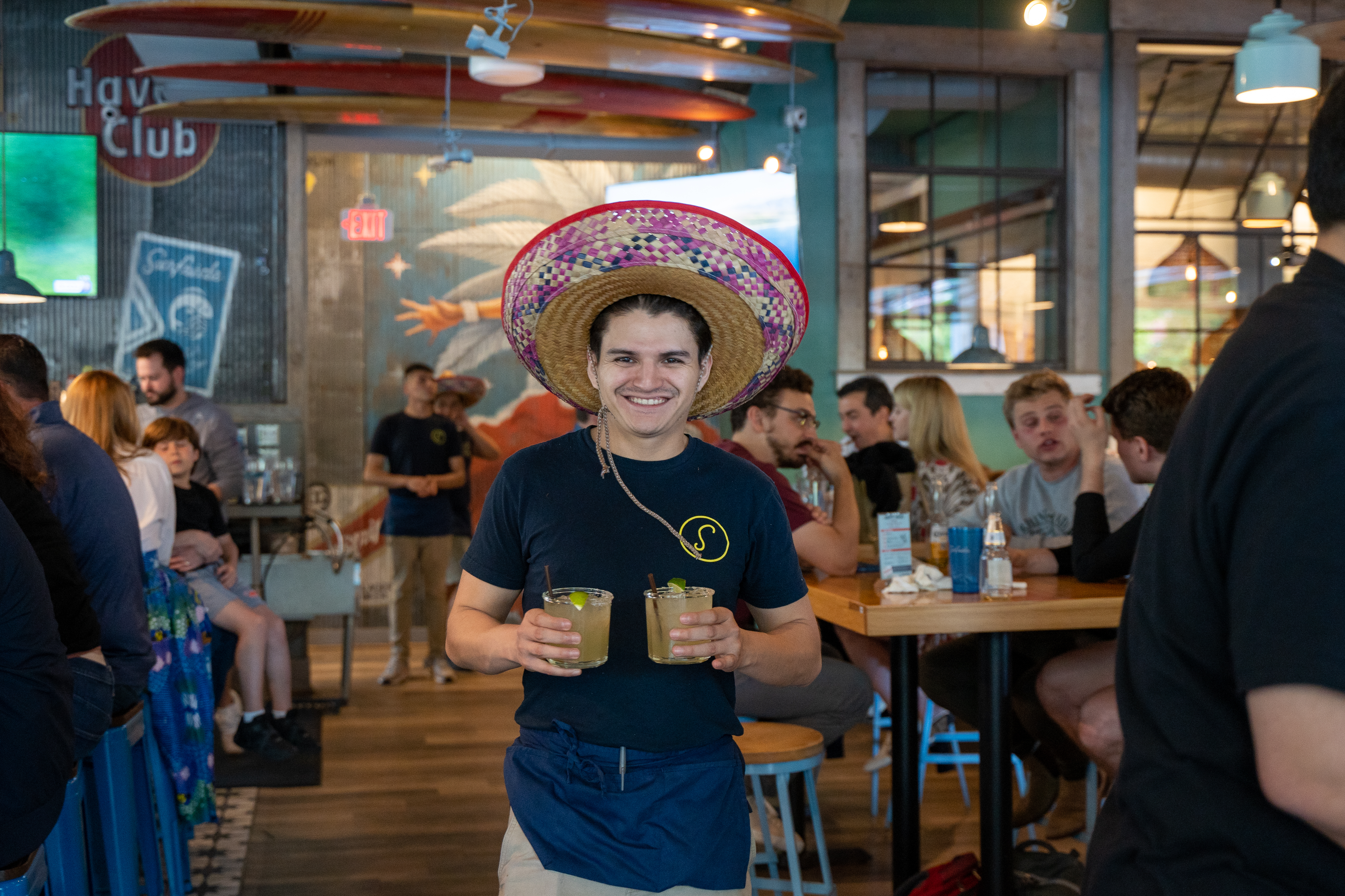 a smiling man holding two glasses and wearing a hat
