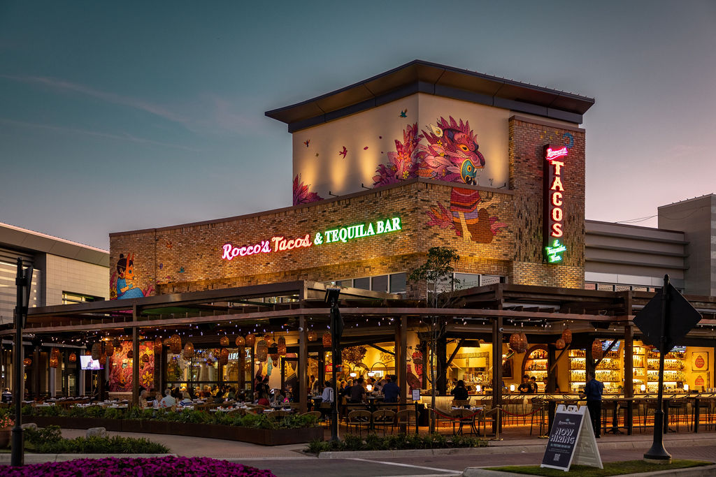 Rocco's Tacos & Tequila Bar exterior at dusk with its neon Tacos sign and covered patio at University Town Center, Sarasota