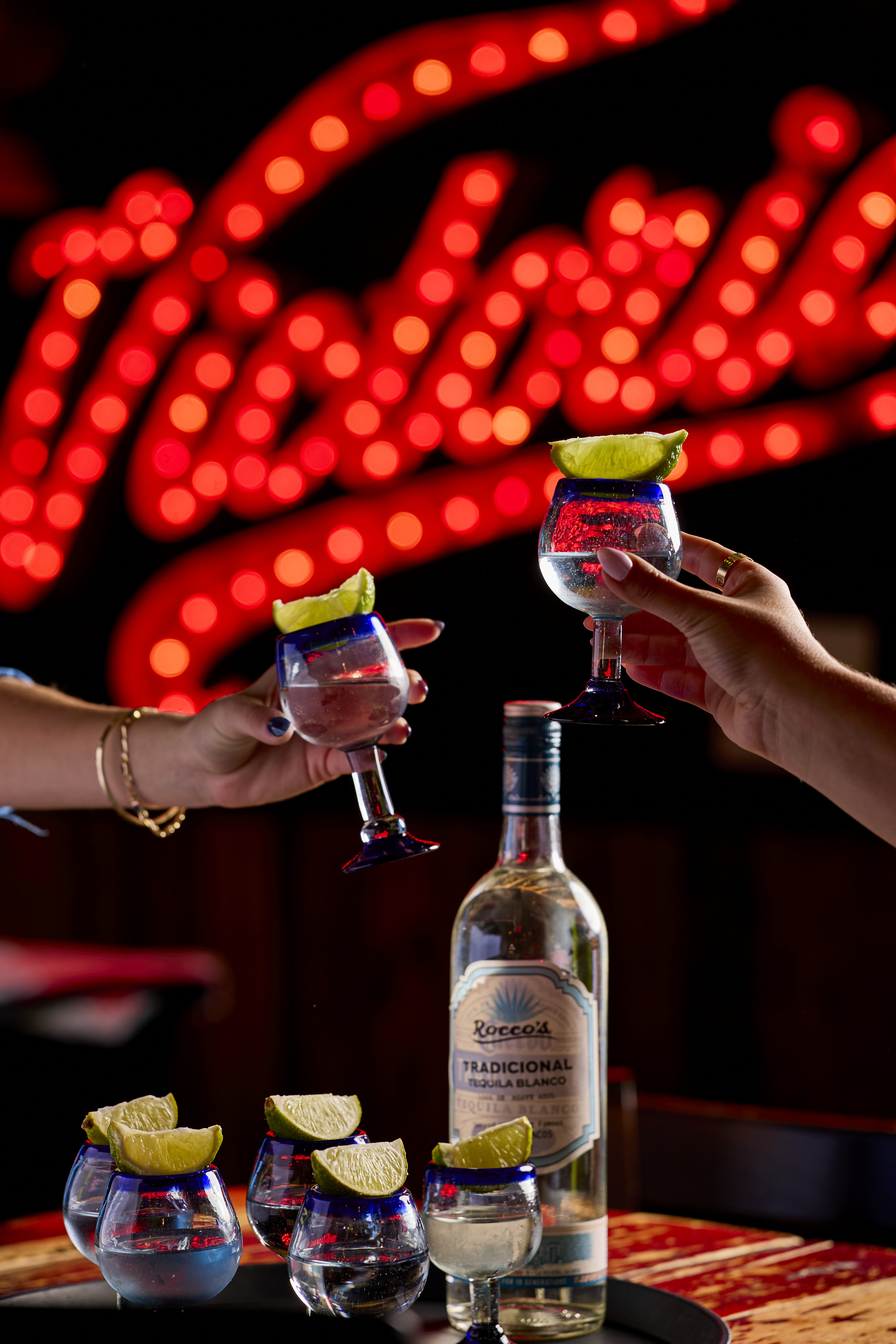 Two guests clinking tequila glasses over a bottle of Tradicional silver tequila and lime wedges at the Rocco's Tacos bar