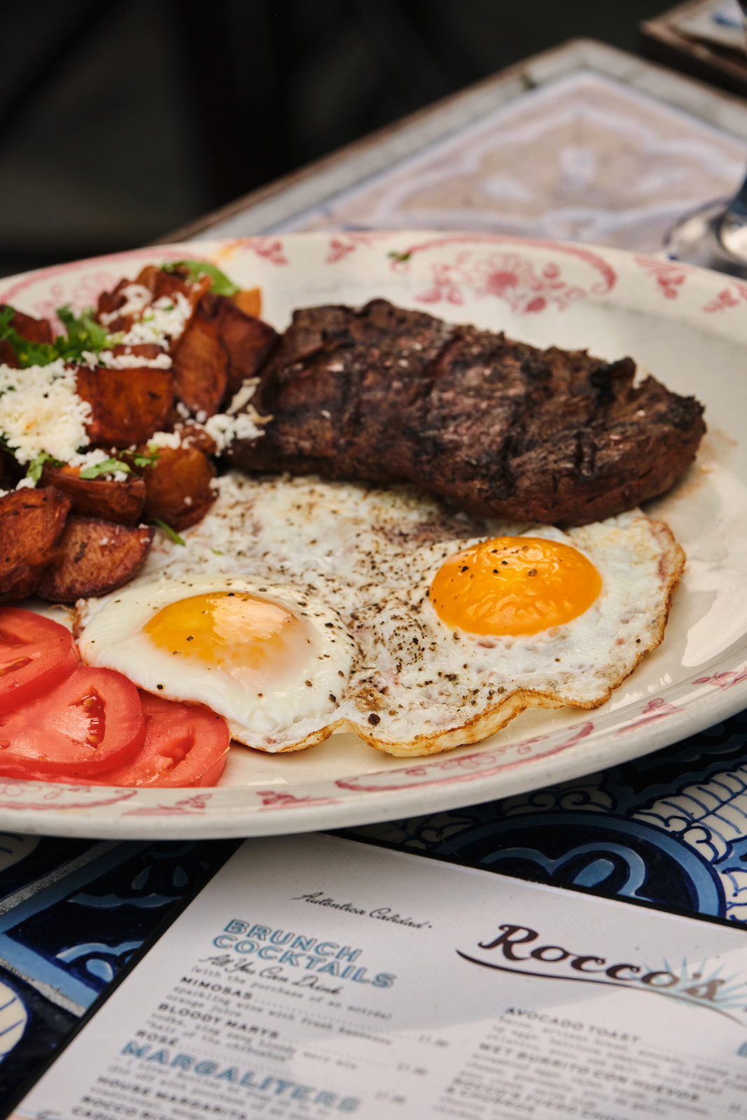 Steak and eggs brunch plate with sunny-side-up eggs, breakfast potatoes and tomato beside the brunch cocktail menu at Rocco's Tacos on Las Olas