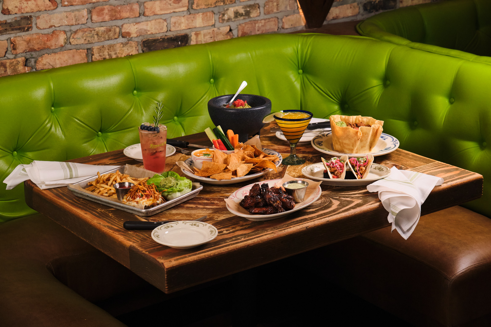 Molcajete guacamole, tacos, chips and cocktails spread across a table in a green booth at Rocco's Tacos at Mercato in Naples