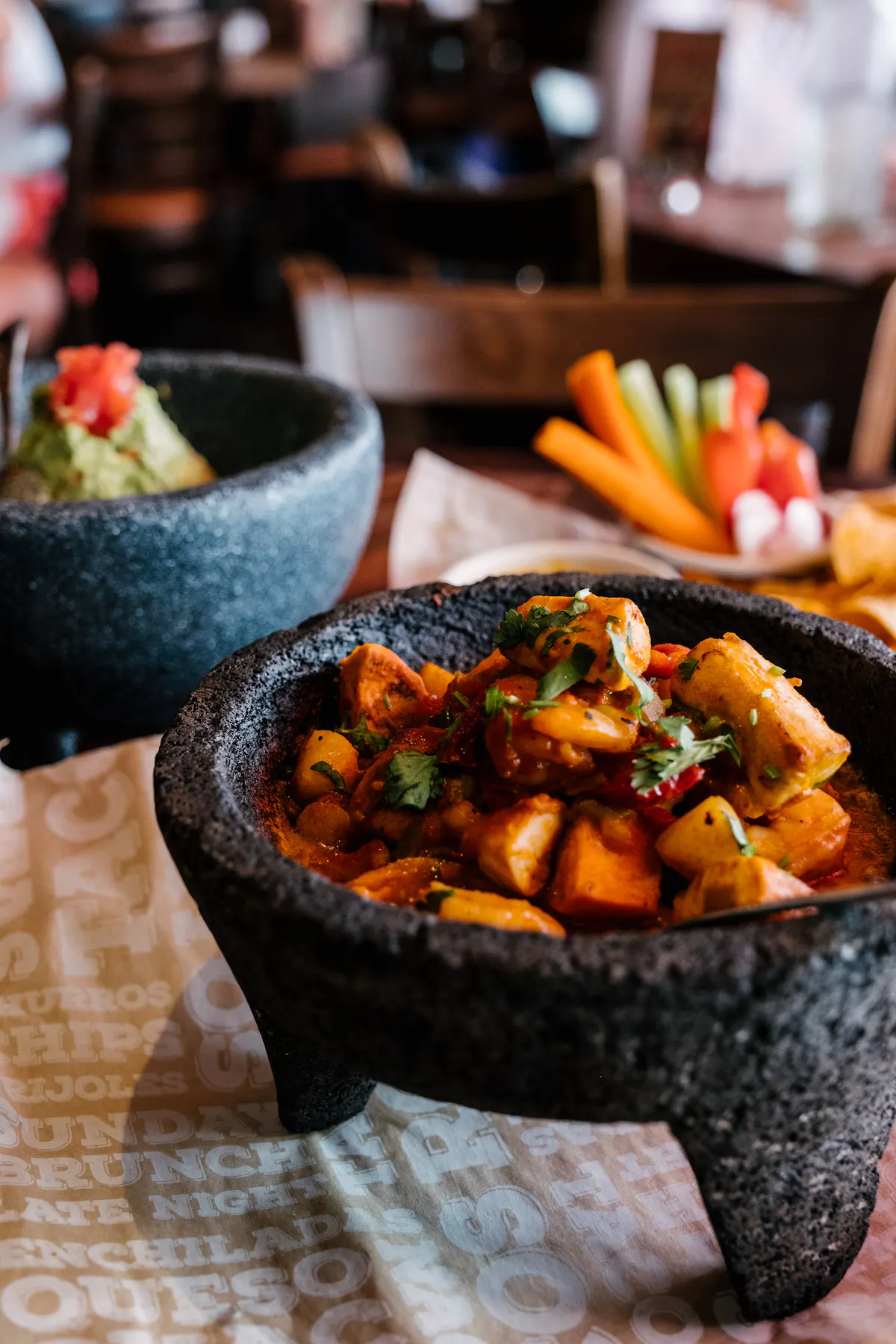 Sizzling Molcajete entrée in a volcanic-rock bowl at Rocco's Tacos, with tableside guacamole and fresh-cut veggies behind it
