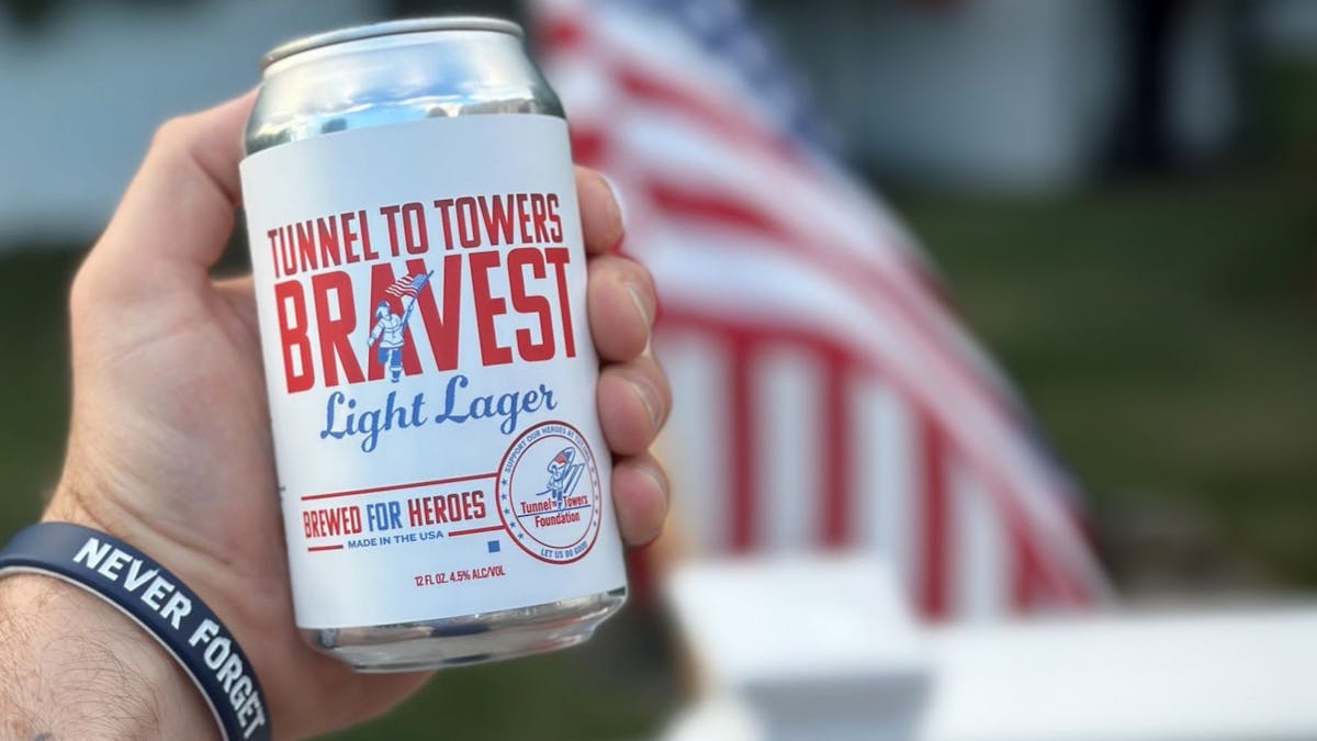 a close up of a person holding a can of Flagship's Bravest Light Lager wearing a bracelet that says Never Forget and a US flag in the background