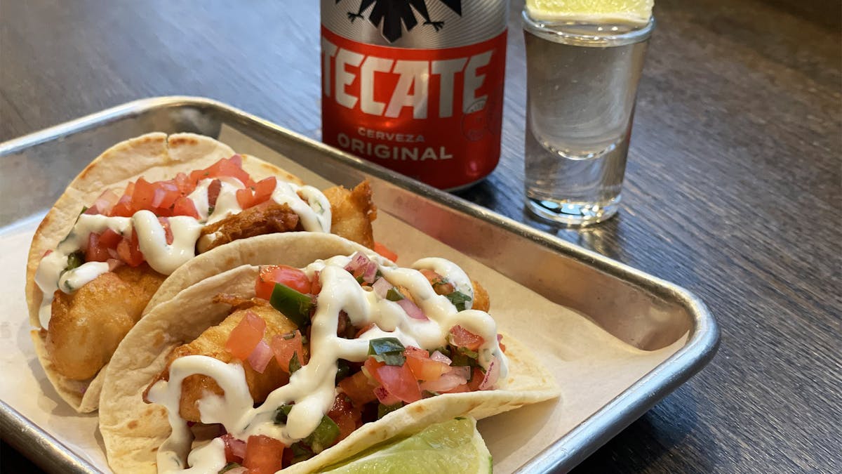 two Baja Fish tacos on a tray with beer can and tequila shot for $16