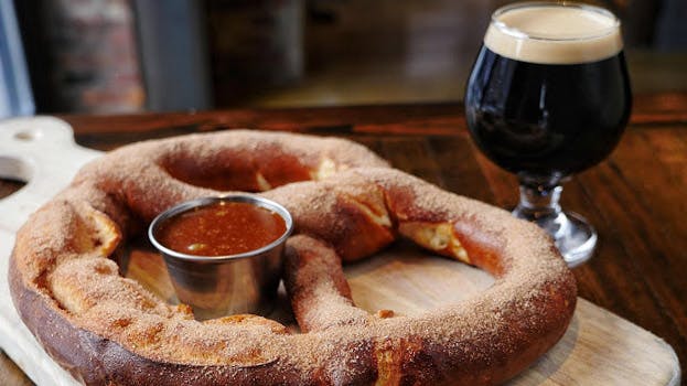 a close up of a pretzel sitting on top of a wooden cutting board and a glass of stout