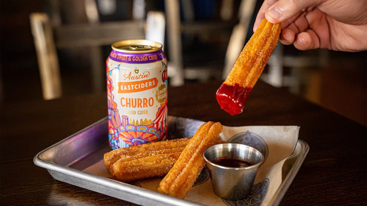 hand dipping churro into raspberry sauce with Austin Eastciders Churro Cider can in the back