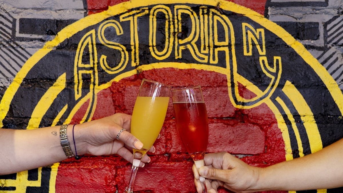 two hands cheersing with mimosas against the painted wall that says Astoria NY