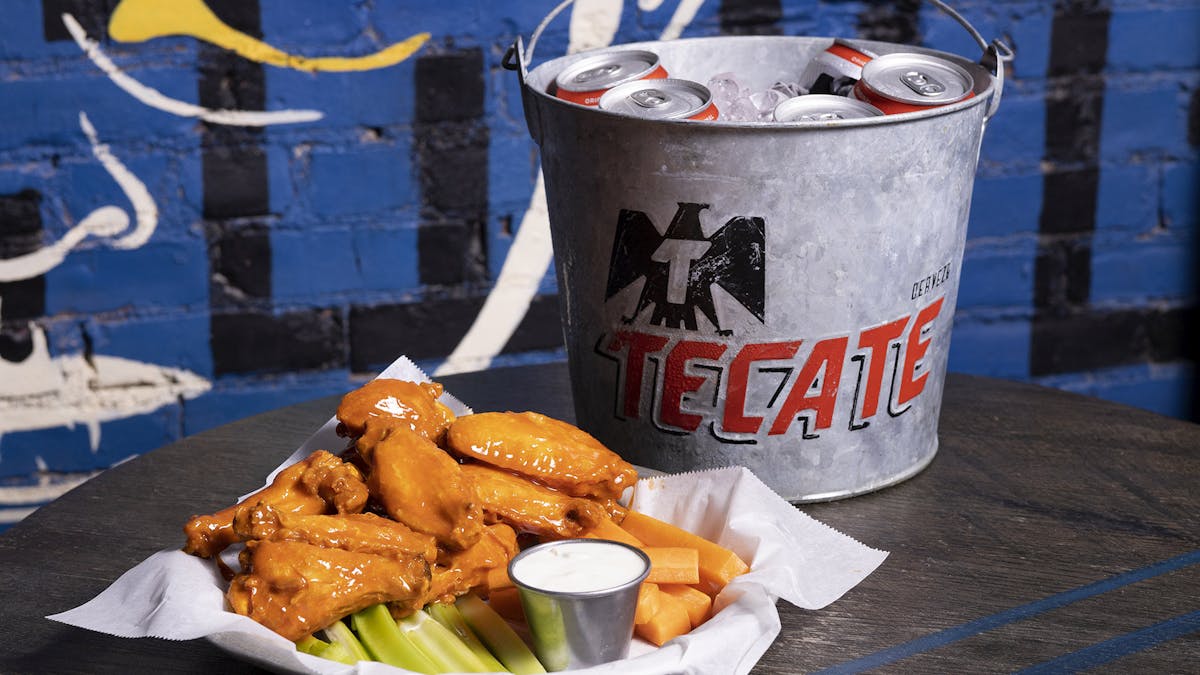 a tray of Buffalo wings with blue cheese dressing and a bucket of Tecate beer on a wooden table