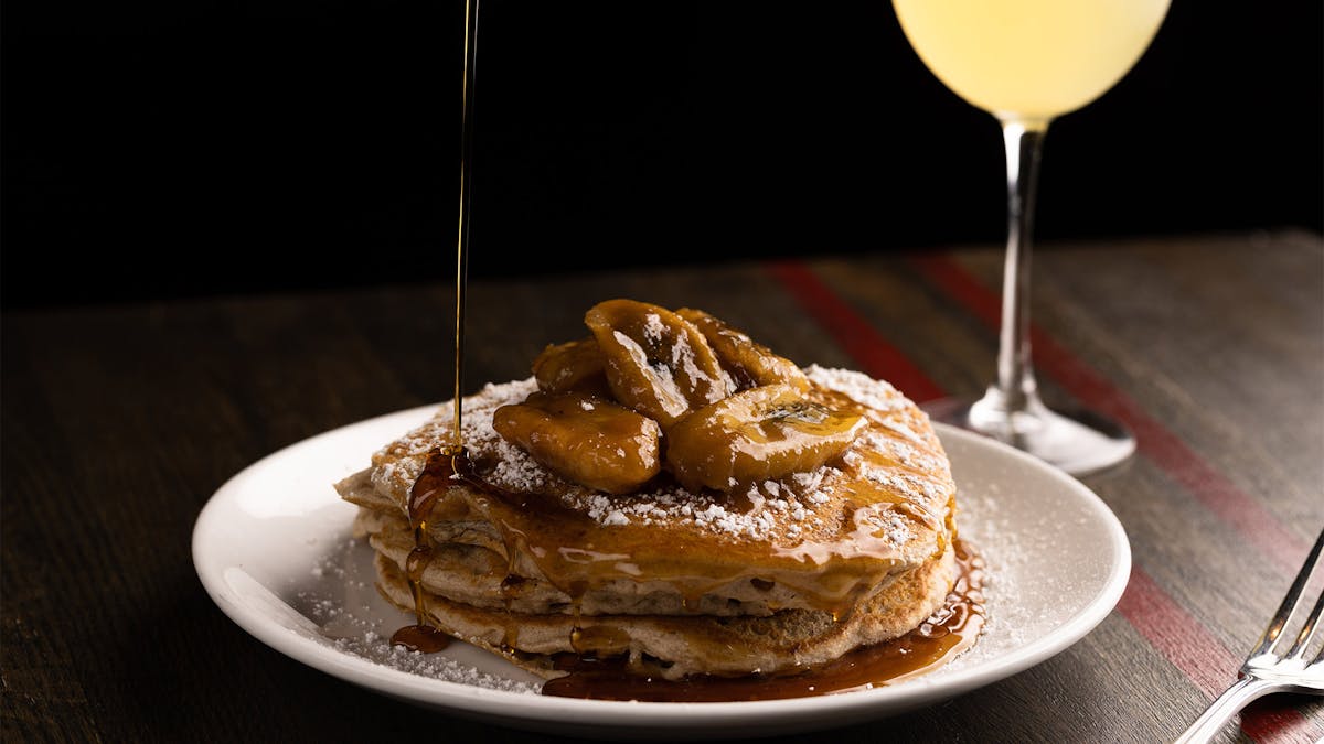 Vegan Bananas Foster Pancakes being poured with maple syrup on a wooden table and a glass of mimosa in the back