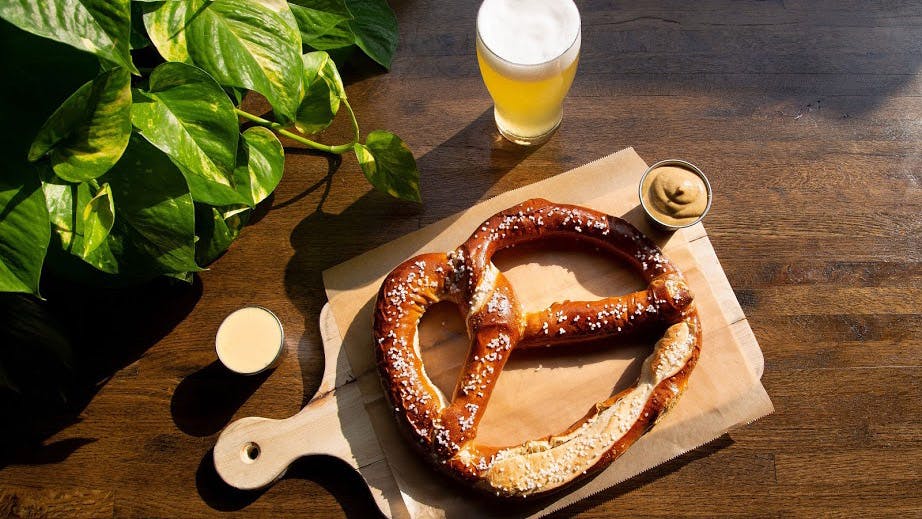 a pretzel sitting on top of a wooden board with mustard sauce on the side and beer glass with a plant in the corner